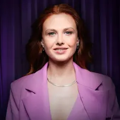 Anna Karczmarczyk w różowej marynarce, z rudymi włosami i niebieskimi oczami, uśmiecha się na tle fioletowej kurtyny. Jej filmy, seriale i programy cieszą się popularnością.