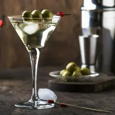 Elegancki drink z Martini Bianco, ozdobiony oliwkami, z kostkami lodu i akcesoriami barmańskimi w tle. Idealny przepis na to, jak zrobić drinka z martini bianco.