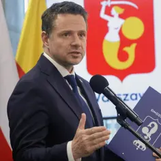 Prezydent Warszawy w garniturze przemawia przy mikrofonie, trzymając teczkę z herbem miasta.