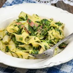 Pappardelle z boczkiem i szpinakiem, podane na białym talerzu, z widelcem.