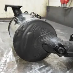 Część układu wydechowego, prawdopodobnie tłumik fap, owinięty czarną taśmą, leży na metalowym stole.