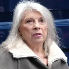 Kobieta w jesiennym wieku, z siwymi włosami i w zielonym płaszczu, patrzy wprost.