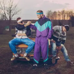 Trzech mężczyzn w strojach z lat 90. moda, siedzi na pomarańczowym samochodzie. Jeden ściska dłoń drugiemu.