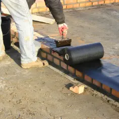 Pracownik nakłada masę asfaltowo kauczukową na mur ceglany, przygotowując podkład pod hydroizolację.