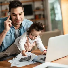 Ojciec rozmawia przez telefon, trzymając dziecko, które dotyka laptopa. Obok leży kalkulator i dokumenty. Zastanawia się, ile to netto z 4700 brutto.