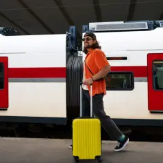 Mężczyzna z żółtą walizką na kółkach idzie obok pociągu z czerwonymi drzwiami.