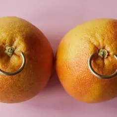 Dwa grejpfruty z metalowymi kółkami w miejscu, gdzie mogłyby być sutki, przypominające nipple piercing.