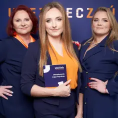 Trzy kobiety w granatowych marynarkach i pomarańczowych bluzkach stoją przed niebieską ścianą z logo.