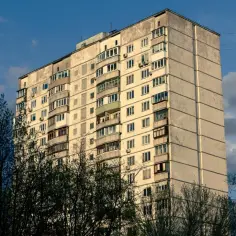 Blok mieszkalny z balkonami, otoczony drzewami. W tle błękitne niebo z chmurami. Może to miejsce, gdzie ktoś ma służebność mieszkania.