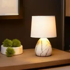 Elegancka lampa ceramiczna stołowa z marmurowym wzorem, oświetlająca drewniany stolik obok zielonych dekoracji.