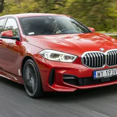 Czerwone BMW F40 w ruchu, z widocznym kierowcą za kierownicą, na tle rozmytego krajobrazu.
