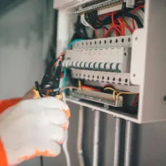 Elektryk w białych rękawicach pracuje przy skrzynce z bezpiecznikami. Zabezpieczenie przedlicznikowe jest kluczowe dla bezpieczeństwa instalacji.