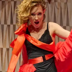 Aktorka w czerwonej sukni i czarnym gorsecie na scenie mniejszej Teatru Capitol, z burzą blond włosów i otwartymi ustami.
