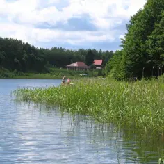 Dwie osoby stoją nad brzegiem jeziora Gim, otoczonego zielenią. W oddali widać domy.