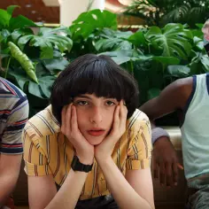 Trzech chłopców z Stranger Things 3 siedzi obok siebie, otoczonych bujną zielenią.