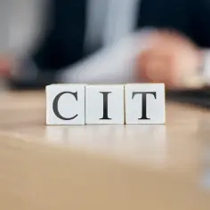 Trzy białe kostki z literami C, I, T tworzą napis CIT. W tle rozmyta postać osoby przy biurku, symbolizująca rozliczenia podatkowe.
