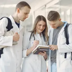 Trzech studentów medycyny w kitlach dyskutuje nad książkami. Po analityce medycznej można rozwijać karierę naukową lub pracować w diagnostyce.