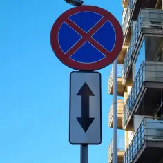 Znak zakazu parkowania przed budynkiem z balkonami. Strzałki wskazują, że zakaz obowiązuje po obu stronach znaku.