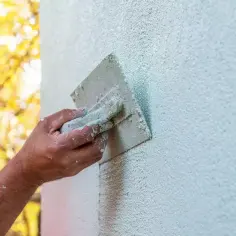 Dłoń zacierająca tynki mineralne na jasnej ścianie. W tle jesienne drzewa.