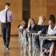 Nauczyciel obserwuje uczniów piszących egzamin w szkolnych ławkach.