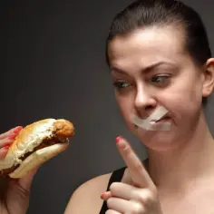 Kobieta z zaklejoną taśmą ustami patrzy na burgera, walcząc z odczuwaniem głodu.
