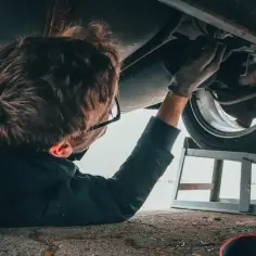 Mechanik bada podwozie auta, sprawdzając, jak sprawdzić czy prądnica jest sprawna. W tle puszka WD-40 i podnośnik.