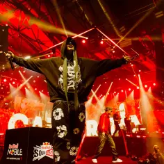 Raper w kapturze i ozdobnych spodniach na scenie podczas Red Bull Soundclash, z mikrofonem w dłoni i okularami w drugiej.