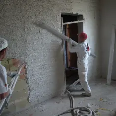 Dwóch budowlańców w białych kombinezonach nakłada tynki w workach na ściany. Jeden z nich używa długiej łopaty.
