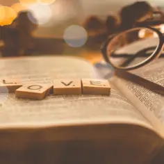 Drewniane klocki z literami "LOVE" na otwartej księdze, symbolizujące przykazanie miłości. Okulary leżą obok.