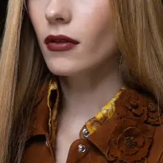 Kobieta z długimi blond włosami i ustami w kolorze karmelowym, ubrana w brązową kurtkę z zamszu ozdobioną kwiatami.