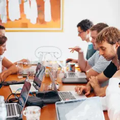 Grupa ludzi pracuje przy stołach z laptopami. To miejsce, gdzie może pracować socjolog, analizując dane i tworząc raporty.