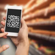 Dłoń trzyma smartfon z widoczną na ekranie aplikacją do skanowania kodów QR. Tło sugeruje sklep.