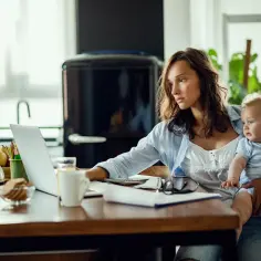 Kobieta z dzieckiem na rękach pracuje przy laptopie. Zastanawia się, kto wypłaca macierzyński po ustaniu zatrudnienia, szukając odpowiedzi w internecie.