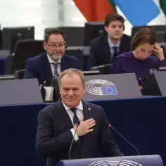 Donald Tusk przemawia na forum, z dłonią na sercu. W tle siedzą posłowie, tworząc trzeci rząd Donalda Tuska.