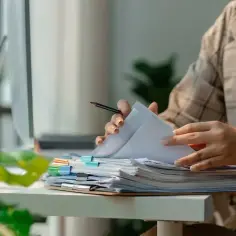 Pracownik przegląda dokumenty, marząc o urlopie dla poratowania zdrowia. Stos papierów czeka na biurku, a za oknem zieleń.