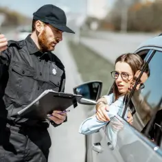 Policjant w czapce i mundurze rozmawia z kobietą w samochodzie, trzymając teczkę. Może to być początek drogi, jak zostać komisarzem policji.