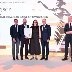 Pięć osób w garniturach i sukience pozuje do zdjęcia z dyplomem na scenie.