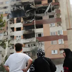 Zniszczony budynek mieszkalny, ludzie patrzą na pożar, w tle drabina strażacka.