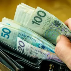 Dłoń wsuwa banknot 100 zł do portfela z innymi banknotami. To symbol oszczędzania, być może na obligacje indeksowane inflacją.