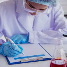 Osoba w fartuchu laboratoryjnym i rękawiczkach pisze na kartce obok mikroskopu i probówek.