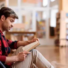 Młody mężczyzna w kratę czyta książkę w bibliotece. Wierzy, że wiedza i pasja sprawią, że "non omnis moriar".