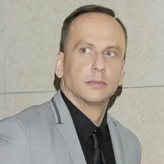 Dariusz Krupa obecnie, w szarym garniturze i czarnej koszuli, spogląda w bok.