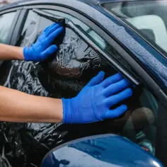 Ręce w niebieskich rękawiczkach odklejają czarną folię z szyby samochodu. Pokazano, jak zdjąć folię z szyby.