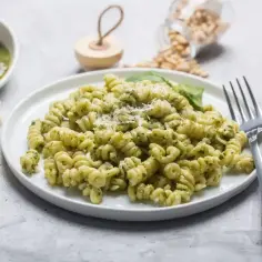 Talerz makaronu fusilli z pesto, posypany parmezanem, obok kawałki sera i miseczka sosu.