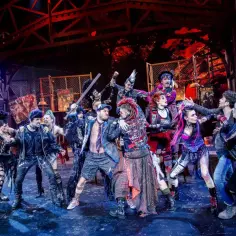 Grupa punkowych postaci na scenie, w tym z irokezami i w skórzanych kurtkach, w dynamicznej scenie z musicalu "We Will Rock You".