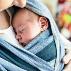 Śpiące niemowlę otulone niebieskim otulaczem, blisko twarzy opiekuna. Może to być przykład źle noszenia niemowlaka, jeśli otulacz nie jest odpowiednio zawiązany.