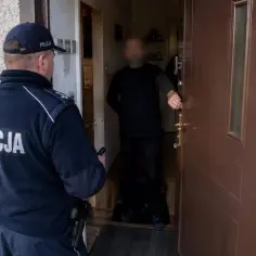 Policjant w mundurze stoi przed drzwiami, gdzie inny mężczyzna otwiera je. Sytuacja sugeruje naruszenie miru domowego.