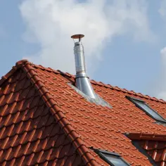 Metalowy komin na czerwonym, dachówkowym dachu pod błękitnym niebem z chmurami.