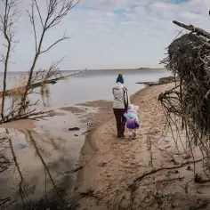 Kobieta z dzieckiem spaceruje po piaszczystym brzegu przy ujściu rzeki do morza.