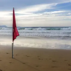 Czerwono-żółta flaga ostrzega przed niebezpieczeństwem na plaży. Surferzy korzystają z fal, podczas gdy na piasku widać ślady stóp.